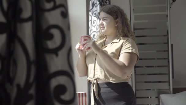 Sonriente encantadora mujer caucásica bebiendo café por la mañana y mirando por la ventana. Retrato de vista lateral de la señora feliz con sobrepeso disfrutando de una bebida caliente en casa. Felicidad y estilo de vida. — Vídeos de Stock