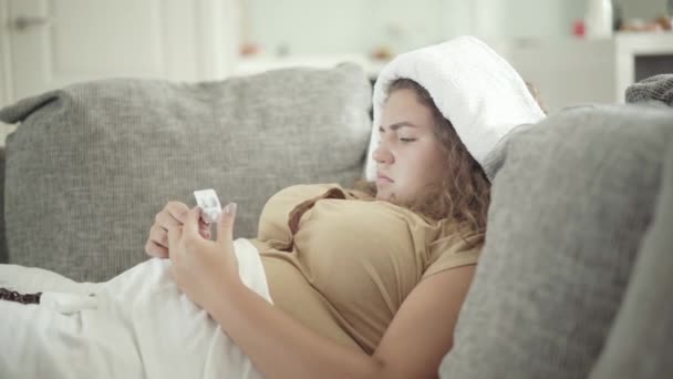 Koncentrerad ung fyllig kvinna väljer piller liggande på soffan med handduk på huvudet. Sidovy porträtt av sjuka kaukasiska plus storlek dam med förkylning och influensa, feber, och hög temperatur. — Stockvideo