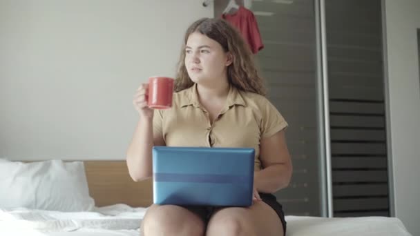 Egy magabiztos fiatal nő, aki reggel e-maileket néz és kávét iszik. Pozitív fehér kövér nő portréja, aki az ágyon ül és gépel a laptop billentyűzetén. Életmód-koncepció. — Stock videók