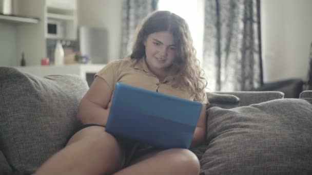 Portrait of cheerful plump woman using social media on laptop and smiling. Young overweight Caucasian lady sitting on couch and surfing Internet indoors. Lifestyle and wireless communication. — Stock Video