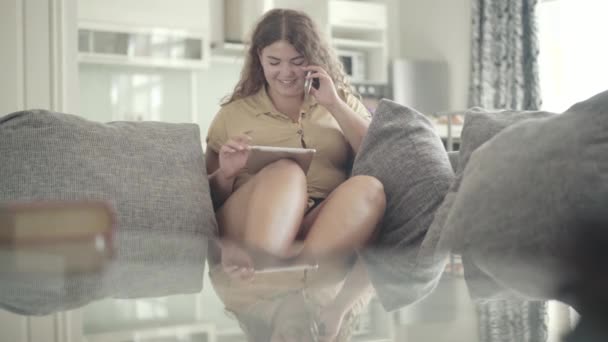 可爱的加尺码的黑发女人在平板电脑上上网和打电话。白种人年轻的卷发女士躺在沙发上，面带微笑的全景画像. — 图库视频影像