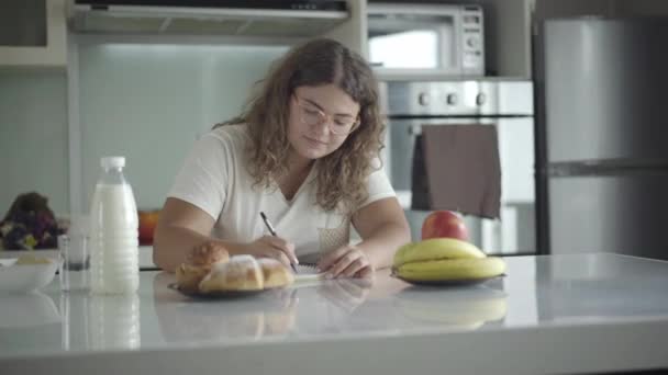 Jeune femme obèse caucasienne calculer les calories assis dans la cuisine le matin. Portrait de dame concentrée dans les lunettes au choix entre pomme saine et croissant délicieux frais. — Video