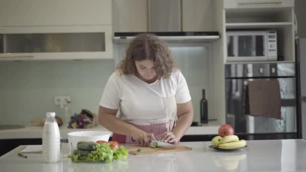 Mulher obesa caucasiana cozinhar salada fresca na cozinha. Retrato de sobrepeso jovem concentrada senhora cortar legumes em casa. Conceito culinário e dietético. — Vídeo de Stock