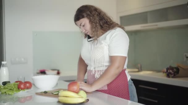 Buclatá žena mluví po telefonu v kuchyni a vaří čerstvý zdravý salát. Portrét mladé, nadměrně velké bělošky, která je ráno doma. Kulinářské a komunikační koncepce. — Stock video