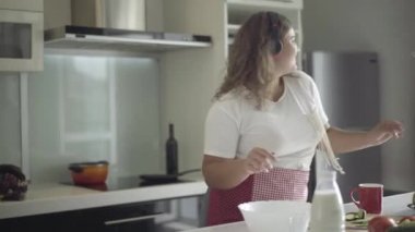 Mutfakta dans eden ve sağlıklı salatalıklar yiyen neşeli, tombul bir kadın. Neşeli, beyaz, obez bir bayanın portresi güneşli bir hafta sonu sabahının keyfini çıkarıyor..