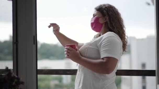 Боковой вид веселой пухлой женщины в розовой маске Ковид-19, стоящей на балконе с кофе и машущей рукой. Портрет радостной молодой кавказки, приветствующей соседей утром во время коронавируса. — стоковое видео