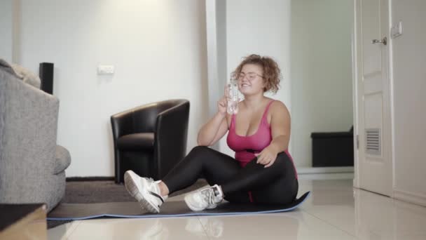 许多穿着运动服的白人女性坐在运动垫上喝着清凉的水。在家训练后正在休息的超重阳性女士的画像. — 图库视频影像
