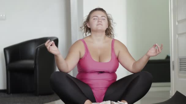 Plan large de sportive joufflue détendue assise dans une pose de lotus les yeux fermés. Portrait d'une femme blanche absorbée faisant du yoga à la maison. Femme en surpoids yogi relaxant à l'intérieur. — Video