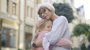 Gün ışığında sakin, sakin, yaşlı bir kadın ve küçük bir kızın sarılışının yan görüntüsü. Şehir caddesinde kucaklaşan mutlu beyaz torun ve büyükannenin portresi..