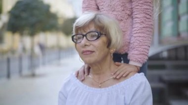 Gözlüklü kendinden emin yaşlı büyükannenin portresi arka planda omuzlarını tutarak duran tanınamaz bir torun gibi görünüyor. Mutlu beyaz kadın güneşli hafta sonunun tadını çıkarıyor..
