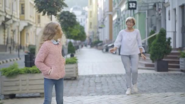 Positief klein meisje loopt op straat met wazig oudere vrouw wandelen op de achtergrond en vertrekken. Portret van vrolijke blanke kleindochter leidend grootmoeder op zonnige dag buiten. — Stockvideo