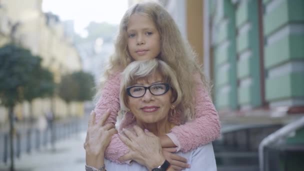 Close-up portret van een gelukkig klein meisje en een oudere vrouw die buiten poseert. Blanke grootmoeder en kleindochter die naar de camera kijken en glimlachen. Geluk en vrije tijd voor het gezin. — Stockvideo