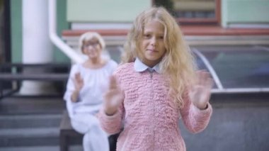 Olumlu bir kızın arka planda oturan bulanık yaşlı bir kadınla el sallayıp onayladığı bir fotoğraf. Güzel beyaz bir torunun güzel portresi. Büyükannemle güneşli bir hafta sonunun keyfini çıkarıyor..