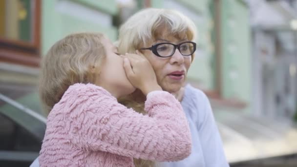Blond liten flicka dela hemligheter med elegant senior kvinna i glasögon. Charmig vit barnbarn viskar på örat av vackra mormor sitter på solig stadsgata. Förtroende och enighet. — Stockvideo