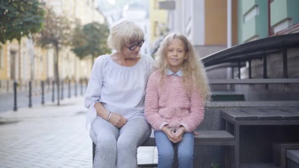 Porträtt av självsäkra vita barnbarn och mormor som korsar ben och ler mot kameran. positiv glad senior kvinna i glasögon och söt liten flicka poserar utomhus. — Stockvideo