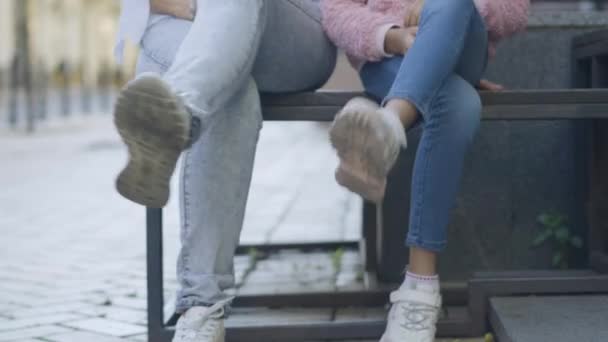 Oigenkännlig kvinna och flicka i avslappnade jeans och sneakers korsar ben sitter på stadens gata. Glad avslappnad mor och dotter eller mormor och barnbarn utomhus. — Stockvideo