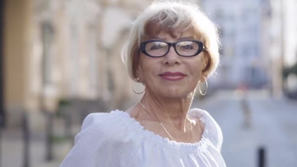 在阳光下，戴着眼镜、面带微笑、自信的老年女性的特写。快乐的白人女性退休人员在城市街道上享受退休金。休闲与幸福. — 图库视频影像