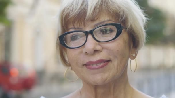 Close-up face of charming senior Caucasian woman in eyeglasses in sunlight outdoors. Portrait of confident beautiful blond lady with brown eyes standing on city street and smiling. — Stock Video