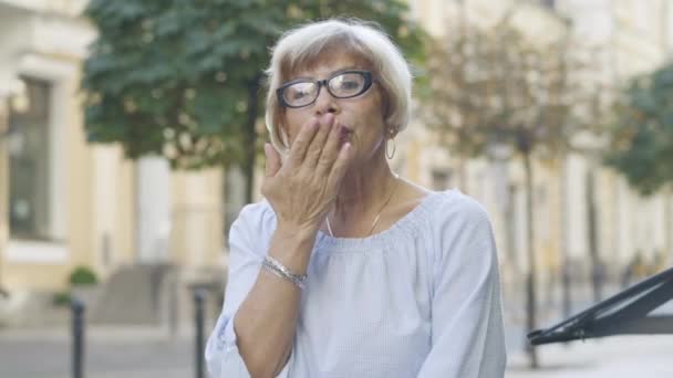 Elegante oudere vrouw in een bril die een luchtkus naar de camera stuurt en glimlacht. Portret van een mooie zelfverzekerde blanke gepensioneerde genietend van een zonnige dag op straat buiten de stad. — Stockvideo
