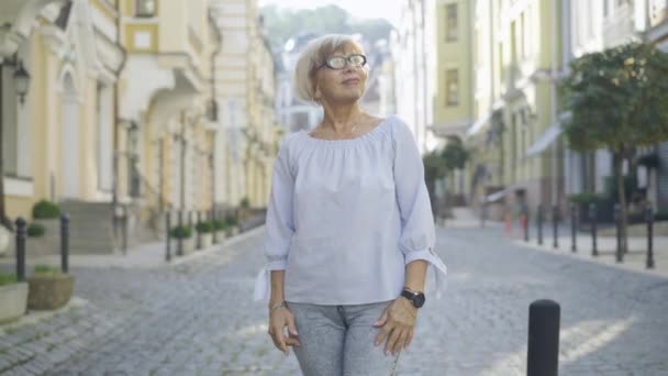 Porträtt av avslappnad kvinnlig pensionär promenader i solljus på stadens gata. Sorglös vit äldre kvinna i glasögon som går utomhus och ler. Glad pensionering och fritid. — Stockvideo