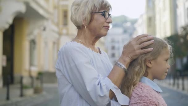 Sidovy av lycklig äldre kvinna i glasögon smeka lockigt hår av söt liten flicka och kyssa barnbarn på pannan. Positiv elegant vit mormor njuter av helgen med barnbarn. — Stockvideo