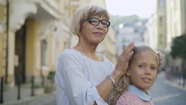 Pewna siebie dziewczynka i starsza kobieta zwracają się do kamery i uśmiechają się, jakby stały na ulicy. Portret zrelaksowanej białej babci i wnuczki na świeżym powietrzu. — Wideo stockowe