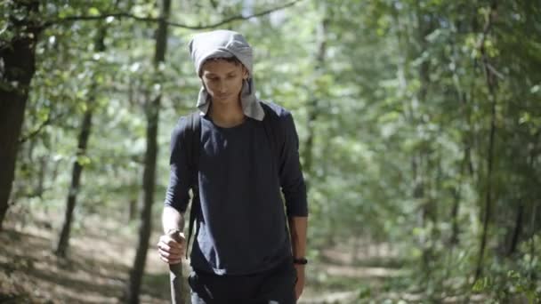 Porträtt av en zigenare som går i solljus i skogen. Ung hemlös man promenader med träpinne utomhus på solig sommardag. Migration och hemlöshet. — Stockvideo