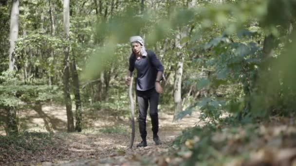 Ampia ripresa di rifugiato maschio esausto che cammina con bastone di legno nella foresta soleggiata. Ritratto di giovane uomo stanco di razza mista che riposa alla luce del sole durante una lunga passeggiata. Concetto di povertà ed emigrazione. — Video Stock