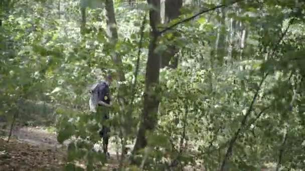 Wide shot side view of young man walking with backpack and wooden stick in sunny summer forest. Refugee or escapee strolling outdoors. Homelessness and migration crisis. — Stock Video