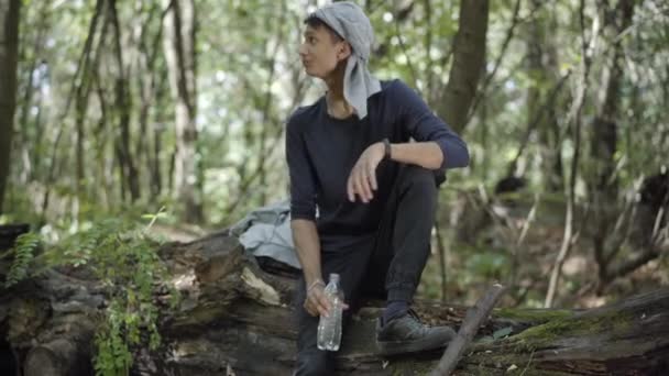 Zenuwachtig bang zigeuner of vluchteling zitten op log in het bos en het drinken van verfrissend water. Portret van een bange jonge gemengde man die zich verschuilt in het bos. Angst en ontsnapping concept. — Stockvideo