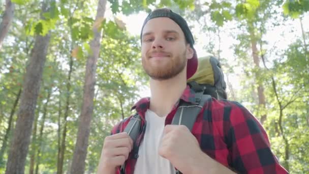 Close-up portrait of confident handsome male hiker standing in sunny forest and smiling at camera. Young Caucasian bearded man posing in sunlight outdoors during adventure travel. Tourism lifestyle. — Stock Video