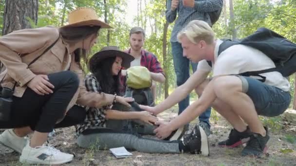 Jovens amigos caucasianos cuidando de mulher com tornozelo ferido. Grupo de turistas na floresta ensolarada de verão dando primeiros socorros e conversando com caminhante triste ter entorse perna. Amizade e apoio. — Vídeo de Stock