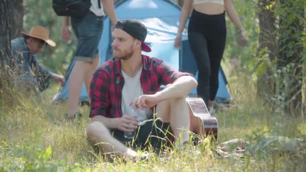 Portret van een ontspannen man met een baard die op groen gras zit en verfrissend water drinkt als wazige vrienden die op de achtergrond een tent opzetten. Zorgeloos Kaukasisch mannelijk toeristisch denken in het zomerwoud. — Stockvideo