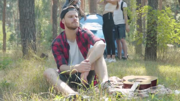 Nachdenklicher junger Mann sitzt auf einer Decke auf einer sonnigen Waldwiese, während Freunde mit Partyhüten zu ihm stoßen. Positive Gruppe gut gelaunter junger Kaukasier feiert Geburtstag auf Wandertour. — Stockvideo