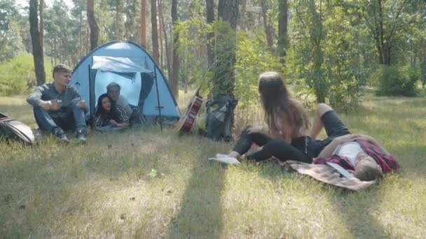 Große Aufnahme fröhlicher junger Freunde, die im Sommerwald plaudern und Tannenzapfen werfen. Eine Gruppe fröhlicher kaukasischer Männer und Frauen zeltet an einem sonnigen Tag im Freien. Freizeit und Freundschaft. — Stockvideo