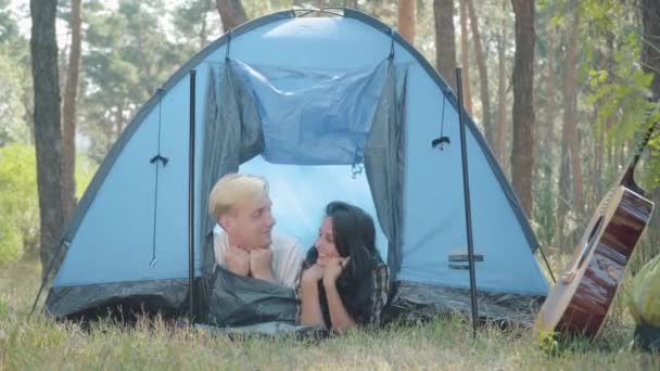Gelukkig jong Kaukasisch echtpaar liggend in een tent in het bos en lachend naar de camera. Portret van een ontspannen knappe blonde man en positieve mooie brunette vrouw kamperen op zomerse dag buiten. Liefde en vreugde. — Stockvideo