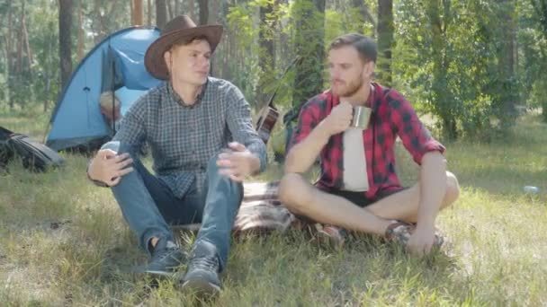 Avslappnade karlar sitter på filt på skogsängen och småpratar. Porträtt av unga stiliga vita män som pratar och dricker te med suddiga vänner i bakgrunden. Camping och livsstil. — Stockvideo