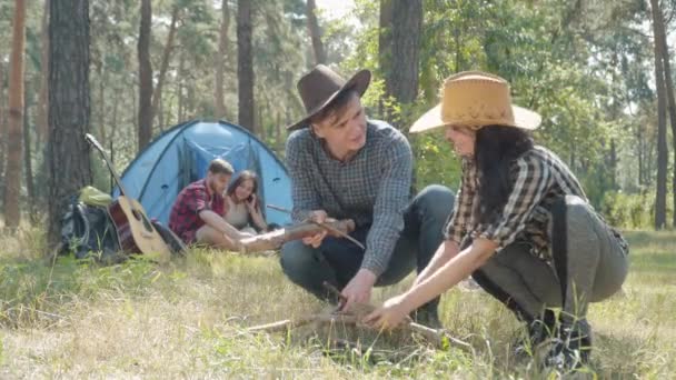 Un jeune homme et une jeune femme positifs mettent le feu dans une forêt ensoleillée d'été avec une tente et des amis campant en arrière-plan. Portrait de heureux détendu touristes caucasiens hommes et femmes en plein air. — Video