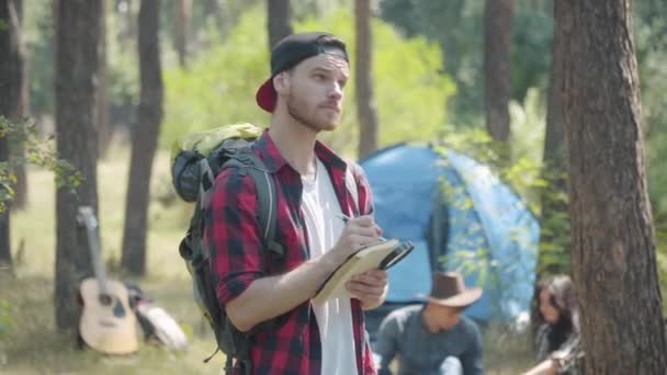 Поглощенный молодой человек смотрит на деревья и пишет в блокноте. Портрет концентрированного кавказского туриста, исследующего природу с группой друзей на заднем плане. — стоковое видео