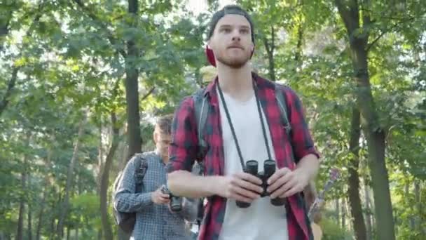 Уверенный мужчина-гид в летнем лесу с группой позитивных молодых туристов, прогуливающихся на заднем плане. Портрет красивого кавказца, идущего и уходящего, как улыбающиеся люди, любующиеся природой. — стоковое видео