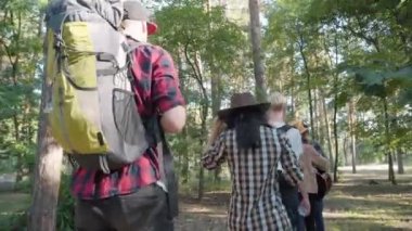 Kamera, yaz günü güneşli orman boyunca sıra sıra yürüyen neşeli genç turistleri izliyor. Rahatlamış erkek ve kadınların, doğa yürüyüşünden zevk aldıklarının arkası..