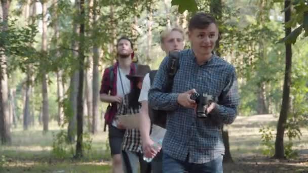 Ряд веселых молодых кавказских мужчин и женщин, прогуливающихся в летнем лесу и уходящих. Один турист останавливается любоваться красотой природы через бинокль и уходит. Друзья, гуляющие на свежем воздухе. — стоковое видео