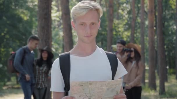 Portrét mladého pohledného blonďatého bělocha pózujícího s papírovou mapou v letním lese s rozmazanými přáteli v pozadí. Sebevědomý muž turista dívá do kamery a usmívá. — Stock video