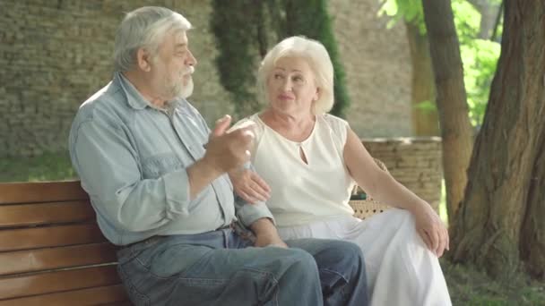 Позитивна старша пара сидить на лавці в сонячному парку і розмовляє. Портрет веселого чоловіка і дружини, які розмовляють і сміються. Щасливі кавказькі пенсіонери із задоволенням зустрічаються на відкритому повітрі.. — стокове відео