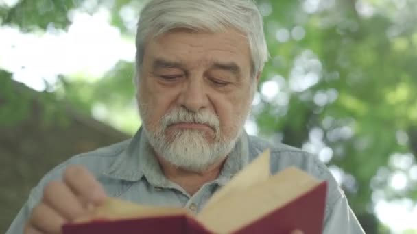 Cara de cerca del viejo barbudo de pelo gris con ojos marrones leyendo libro a la luz del sol al aire libre. Retrato de hombre caucásico absorbido jubilado disfrutando de pasatiempo en el parque de verano. — Vídeo de stock