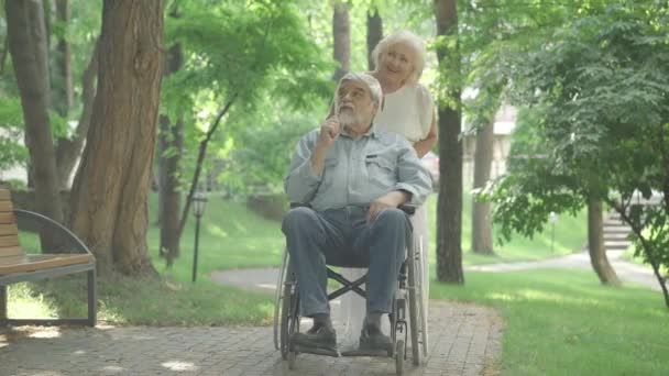 Breed shot van positieve mooie blanke senior vrouw rolstoel met gehandicapte man in het zonlicht buiten. Portret van een liefhebbende vrouw wandelen met verlamde echtgenoot in zomerpark en praten. — Stockvideo
