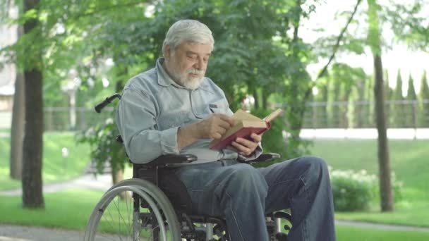 Portrait du vieil homme caucasien en fauteuil roulant lisant le livre à haute voix dans le parc d'été ensoleillé. Un retraité handicapé âgé tourne la page et parle. Loisirs extérieurs de l'homme handicapé. — Video