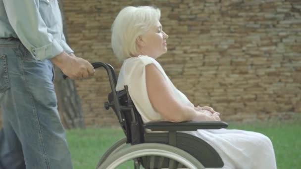 Side view of paraplegic senior woman talking as unrecognizable man rolling wheelchair. Grey-haired Caucasian female retiree on sunny summer day in park with loving husband. Care and support. — Stock Video