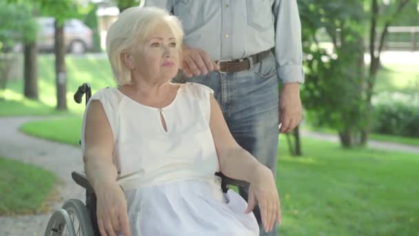 Oigenkännlig man som lägger handen på axeln på en sorgsen förlamad kvinna som sitter i rullstol. Porträtt av deprimerad vit vacker pensionär vilar i solig park med make. Frustrationskoncept. — Stockvideo