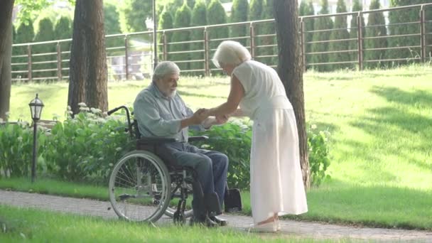 Opiekuńcza starsza kobieta pomaga niepełnosprawnemu mężczyźnie wstać z wózka inwalidzkiego i chodzić. Wdzięczny, biały mąż przytulający kochającą żonę i uśmiechający się. Rehabilitacja i wsparcie. — Wideo stockowe